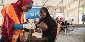 On 17th November 2016, UNICEF Nutrition Officer, Aishat Abdullahi, assesses 7 month old Umara Bukar for malnutrition at a UNICEF supported health clinic at Muna Garage IDP camp, Maiduguri, Borno State, northeast Nigeria as Umaras mother (in black) looks on.  20 days ago Umara weighed just 4.2kg when he first arrived at the health clinic run in partnership with the Nigerian government.  He now weighs 5.1kg.
To date, over 117,00 children with severe acute malnutrition (SAM) in northeast Nigeria have been admitted to therapeutic feeding programmes run by UNICEF and partners.

In November 2016, Fanna Mohammed is worried about her son. 7-month old Umara is thin and listless. He rests his head against his mother's shoulder as she carries him in her arms. The family fled their village in a rural areas of Borno state earlier this year due to the ongoing Boko Haram-related crisis.  Now they live in an IDP camp in the state capital, Maiduguri. Muna Garage IDP camp is home to an estimated 20,000 people. 8,000 of those are children under the age of 5.  Fanna says Umara's been ill for the past few week. He is physically showing signs of severe acute malnutrition. The bones on his chest and back are prominent; his skin is loose around his arms and legs. Fanna's brought Umara to a UNICEF-supported medical clinic that's situated within the camp. Umara is immediately given life-saving treatment. Ready-to-Use Therapeutic Food (RUFT) is a peanut-based paste that's high in calories and full of added vitamins and minerals. Three packets a day, for eight weeks can save the life of a child with severe acute malnutrition. Lying in his mother's arms, Umara slowly but surely eats the food. Before he's allowed home, the health team put him on a course of extra vitamin A, antibiotics and anti-malarial medicine.  A week later, Umara's back at the clinic for his second screening. There's a small, but steady improvement. His arm is plumper, at 9.7CM and he's put on a little weight. The
