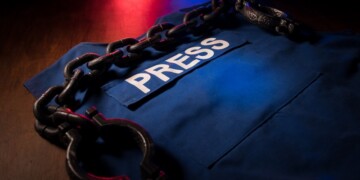 Freedom of the press and journalism concept. Blue journalist (press) vest in dark with backlight and fog. Shackles on vest. Selective focus