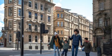 A multiracial family of four, a mother, father and their two little boys on a staycation in the city of Newcastle on a sunny winters day. They are wearing casual, winter clothing and accessories. They are walking down a city street, holding hands and swinging one of the boys in the air.