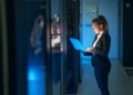 Woman IT specialist in elegant suit working on notebook computer in data center next to server racks