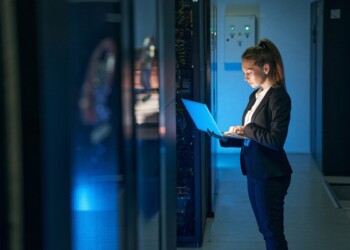 Woman IT specialist in elegant suit working on notebook computer in data center next to server racks