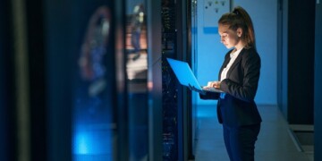 Woman IT specialist in elegant suit working on notebook computer in data center next to server racks