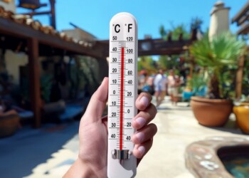Blazing heat in a tropical resort setting. A hand holds a thermometer showing very high temperature, with sunlit courtyard, blurred tourists, palm trees, and summer atmosphere around.