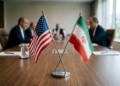 Crossed flags of United States and Iran are displayed on a wooden conference table with blurred officials in background during diplomatic negotiations.