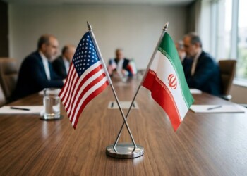 Crossed flags of United States and Iran are displayed on a wooden conference table with blurred officials in background during diplomatic negotiations.